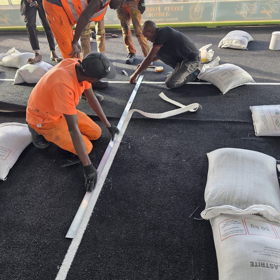 Workers applying precision line markings on the new black padel turf with kiln-dried silica sand infill bags ready